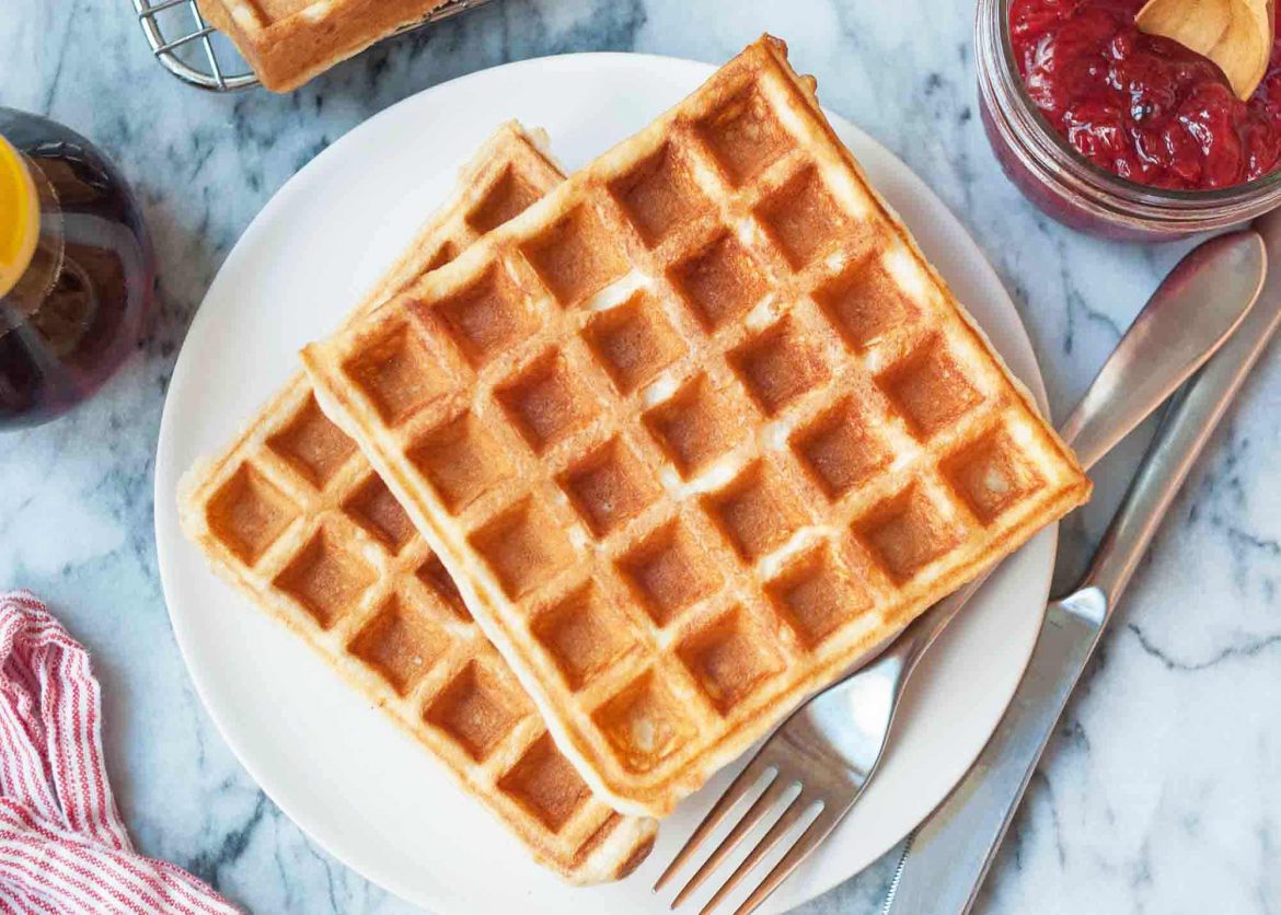 two waffles on a plate with a pair of cutlery, next to a pot of strawberry jam